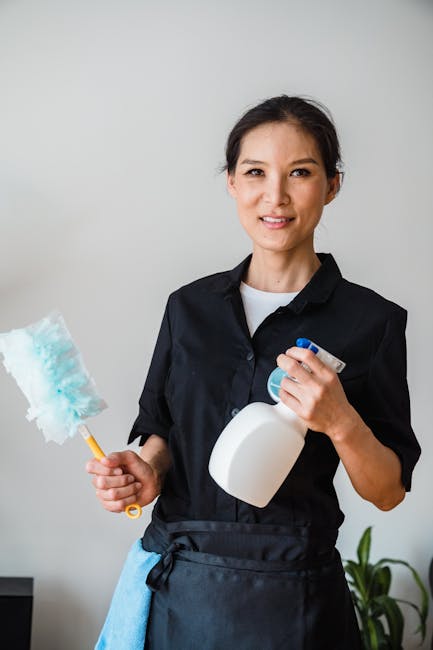A professional cleaner from Cleaners Kennington smiles while holding a cleaning spray bottle in one hand and a blue cleaning cloth attached to a yellow handle in the other. She is wearing a dark unifo