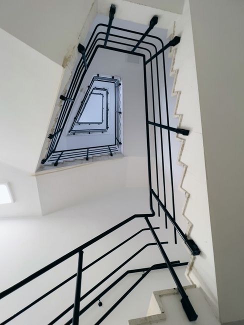 A vertical view looking down a multi-story residential stairwell with a black metal railing and white walls. The staircase features rectangular landings and internal walls painted in a clean, neutral white, illuminated by natural light from an unseen source. The black metal handrail bars are evenly spaced and securely fastened to the walls and stair edges. The stairs are made of concrete or similar material, with visible signs of dust and minimal debris, indicating recent cleaning or maintenance. The overall appearance is tidy and well-maintained, suitable for safe access between floors. This image exemplifies the kind of hygienic, surface-focused deep cleaning and sanitisation services provided by Cleaners Kennington, experts in domestic and communal cleaning solutions for stairwell access problems and improvements.