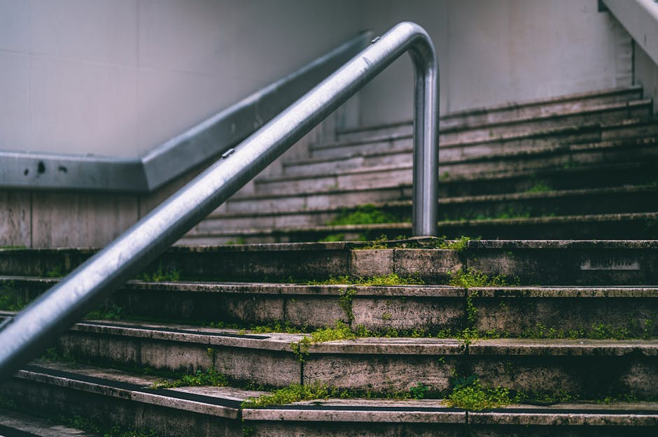 Concrete stairwell outdoors with moss and dirt accumulated on the steps, which are partially covered in algae and grime. A stainless steel handrail runs diagonally across the image, mounted on the side of the stairs, with signs of weathering and corrosion. The lighting is natural, highlighting the rugged surface and the uneven, weathered texture of the concrete steps. The background shows the edge of a building wall, indicating the stairwell's location at a communal or public access point. This image illustrates a common area in need of deep cleaning and maintenance, as part of the services provided by Cleaners Kennington for effective surface cleaning and sanitation.