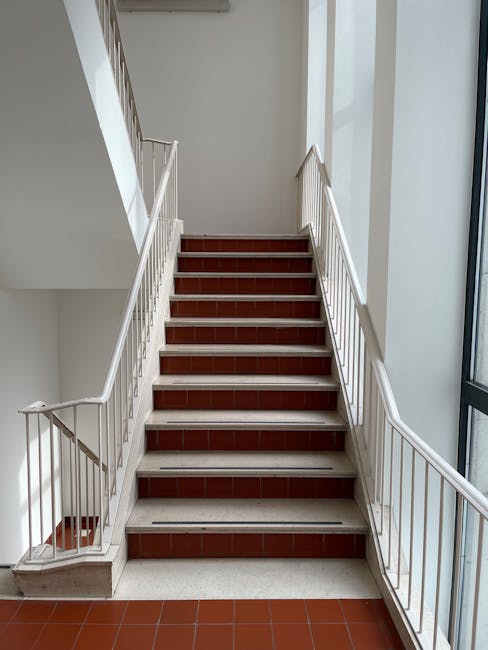 A vertical view looking down a multi-story residential stairwell with a black metal railing and white walls. The staircase features rectangular landings and internal walls painted in a clean, neutral white, illuminated by natural light from an unseen source. The black metal handrail bars are evenly spaced and securely fastened to the walls and stair edges. The stairs are made of concrete or similar material, with visible signs of dust and minimal debris, indicating recent cleaning or maintenance. The overall appearance is tidy and well-maintained, suitable for safe access between floors. This image exemplifies the kind of hygienic, surface-focused deep cleaning and sanitisation services provided by Cleaners Kennington, experts in domestic and communal cleaning solutions for stairwell access problems and improvements.