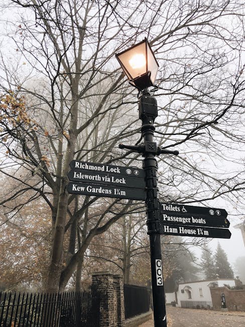 A black metal street lamp with a lit glass lantern standing amidst leafless tree branches and a foggy outdoor setting. Attached to the lamppost are multiple black directional signs with white text, indicating nearby locations such as Richmond Lock, Isleworth via Lock, Kew Gardens 1/4 mile, Toilets & Passenger boats, and Ham House 1/4 mile. In the background, there is a black wrought iron fence, a brick wall, and a few white buildings partially obscured by mist, creating a serene and slightly misty atmosphere typical of a winter day. The lighting is low due to the fog and the lamp's glow, adding a calm and quiet ambiance consistent with an outdoor scene in a park or historic area.