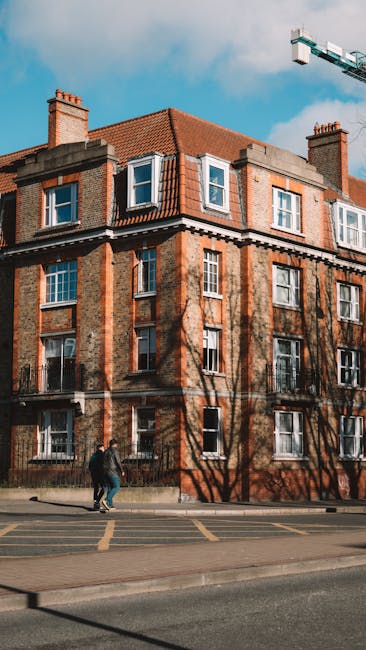 Photograph of a vintage brick residential building with multiple windows, some of which are open, under a partly cloudy blue sky. The building features a red-tiled pitched roof, brick chimney stacks, and external wall details. Shadow patterns from nearby trees are cast onto the facade. In front, two pedestrians are walking along the sidewalk. The image highlights the urban architecture typical of converted flats in Kennington. This scene emphasizes the importance of regular surface cleaning and maintenance for residential exteriors, aligning with the services offered by Cleaners Kennington, as detailed on their website's Kennington Road cleaning checklist for converted flats.