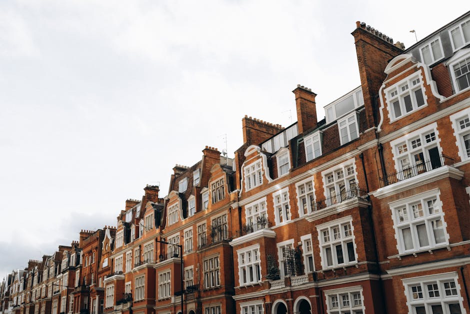 A row of multi-story residential buildings with red brick facades and white decorative window frames. The buildings feature bay windows, small balconies with black railings, and pitched roofs with chimneys. The exterior surfaces appear clean and well-maintained, with natural daylight illuminating the scene under a partly cloudy sky. This image is associated with domestic surface cleaning and deep cleaning services provided by Cleaners Kennington, as mentioned on the Kennington Road cleaning checklist webpage for converted flats.