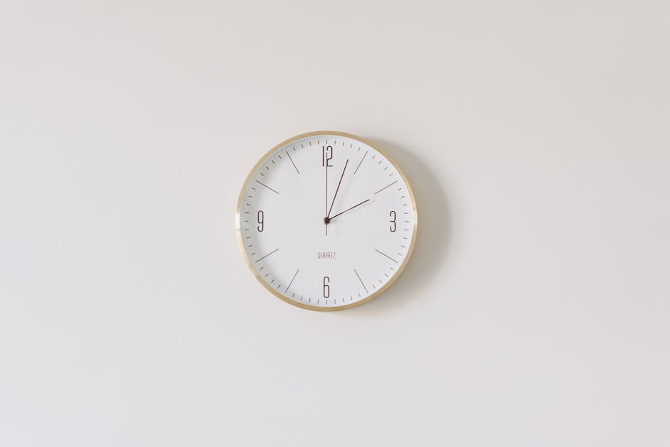 A round wall clock with a white face and a gold rim hangs on a plain, light-colored wall. The clock displays a time just past 12:05, with black hour and minute hands and a thin red second hand. The minimalistic design emphasizes clarity and simplicity. The smooth surface of the wall and the clean appearance of the clock contribute to a sense of tidy, well-maintained space, aligning with the theme of surface cleaning and sanitisation highlighted in the context of last-minute booking fixes with Cleaners Kennington. The soft, even lighting enhances the overall neatness and functionality of the scene, reflecting professional domestic cleaning standards.