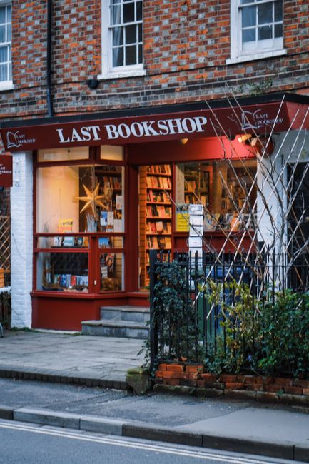 The image depicts the exterior of a bookstore named 'Last Bookshop' located in a brick building with large windows displaying books and a decorative star-shaped light inside. The shop features a brown awning with white text and a small sign for 'Lamps & Lighting' beside the window. In front of the store, there is a paved sidewalk with some steps leading up to the entrance. To the right, a black wrought iron fence surrounds a small area with bushes and trees. The scene is lit with warm interior lighting, contrasting with the cool, dim outdoor lighting, highlighting the clean and well-kept appearance of the storefront, which is relevant to the topic of surface cleaning, sanitisation, and maintenance services offered by Cleaners Kennington, especially in the context of addressing last-minute booking problems and their solutions.