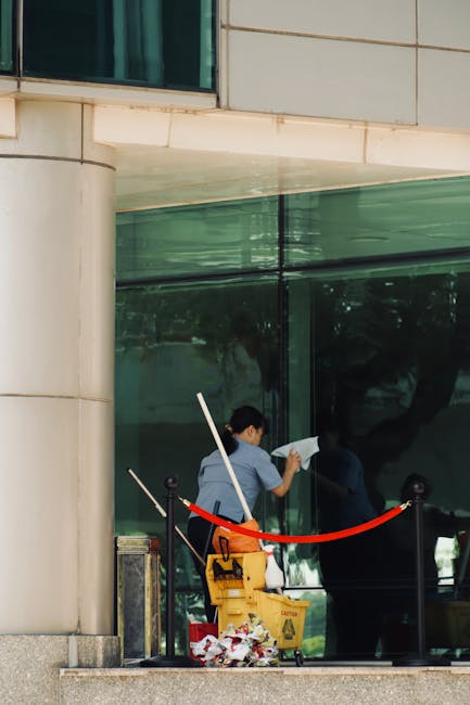 A professional cleaner from Cleaners Kennington is performing surface cleaning on a large glass window of a modern commercial building. The cleaner, dressed in a blue uniform, is using a squeegee or cloth to wipe the glass, ensuring a streak-free, clear finish. The window, framed by beige stone masonry, reflects the surrounding environment, including trees and sky, indicating good natural lighting. A yellow cleaning bucket with cleaning tools, such as a mop handle and cloths, is placed on the ground nearby, along with a caution wet floor sign. The area outside the building features black bollards connected by red ropes, demarcating the cleaning zone. The scene highlights meticulous window sanitisation and deep cleaning processes designed for commercial premises, highlighting the professional standards of Cleaners Kennington for maintaining hygienic, spotless glass surfaces near The Oval, Kennington.