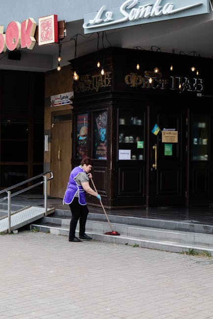 A professional cleaner from Cleaners Kennington is performing surface cleaning on a large glass window of a modern commercial building. The cleaner, dressed in a blue uniform, is using a squeegee or cloth to wipe the glass, ensuring a streak-free, clear finish. The window, framed by beige stone masonry, reflects the surrounding environment, including trees and sky, indicating good natural lighting. A yellow cleaning bucket with cleaning tools, such as a mop handle and cloths, is placed on the ground nearby, along with a caution wet floor sign. The area outside the building features black bollards connected by red ropes, demarcating the cleaning zone. The scene highlights meticulous window sanitisation and deep cleaning processes designed for commercial premises, highlighting the professional standards of Cleaners Kennington for maintaining hygienic, spotless glass surfaces near The Oval, Kennington.