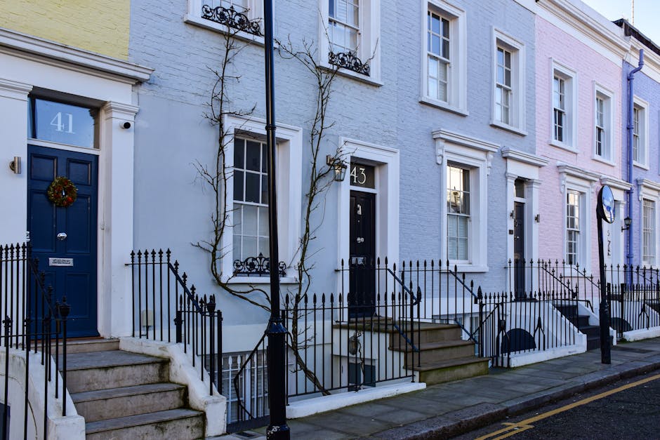 A row of residential houses with painted brick facades in pastel colors, including pale blue, pink, and light purple, each featuring white-framed sash windows and stone steps leading up to the front doors. The front doors are painted in dark blue and black, with decorative doorframes, and some doors have seasonal wreaths. Iron railings line the small front gardens or steps, and black street lamps are positioned along the sidewalk. The street appears clean and well-maintained, with a smooth pavement and minimal litter, under natural daylight conditions highlighting the neat appearance of the properties, suitable for surface cleaning and maintenance guidance by Cleaners Kennington.