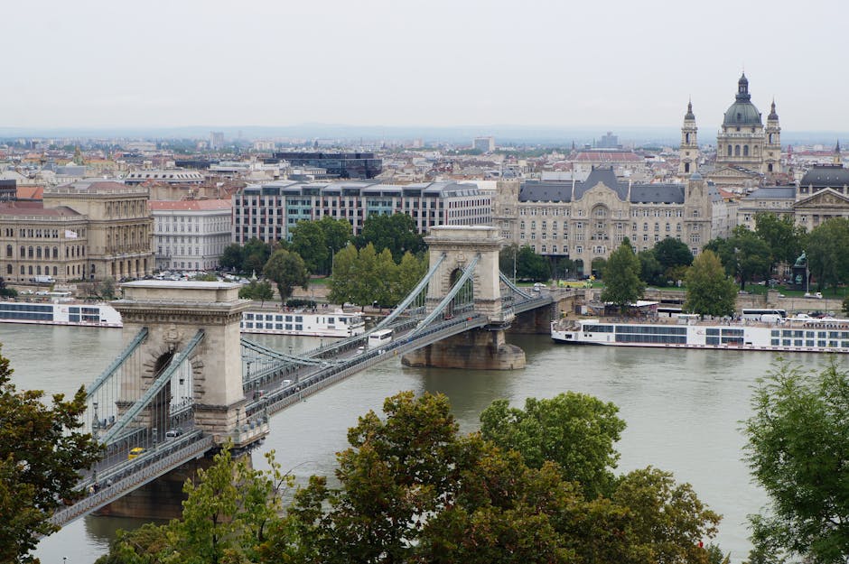 A high-angle view of the Tower Bridge in London with its iconic stone towers and suspension cables spanning the River Thames. The river appears clean with a few boats, including a large white passenger vessel, moored along the sides. Surrounding the bridge are various historic and modern buildings, with some featuring detailed stone facades and others glass windows reflecting the overcast sky. Green trees are visible in the foreground and along the riverbank, adding natural elements to the urban landscape. The scene is evenly lit, showcasing a mix of old and new architecture, with the cityscape extending into the distance under a cloudy sky. This image emphasizes the cleanliness and maintenance of the area, aligning with the themes of surface cleaning and urban sanitation that Cleaners Kennington provides in their residential and commercial cleaning services.