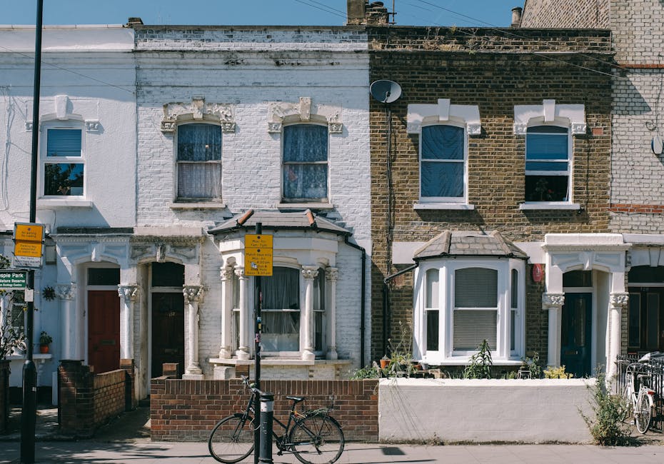 A row of residential houses with painted brick facades in pastel colors, including pale blue, pink, and light purple, each featuring white-framed sash windows and stone steps leading up to the front doors. The front doors are painted in dark blue and black, with decorative doorframes, and some doors have seasonal wreaths. Iron railings line the small front gardens or steps, and black street lamps are positioned along the sidewalk. The street appears clean and well-maintained, with a smooth pavement and minimal litter, under natural daylight conditions highlighting the neat appearance of the properties, suitable for surface cleaning and maintenance guidance by Cleaners Kennington.
