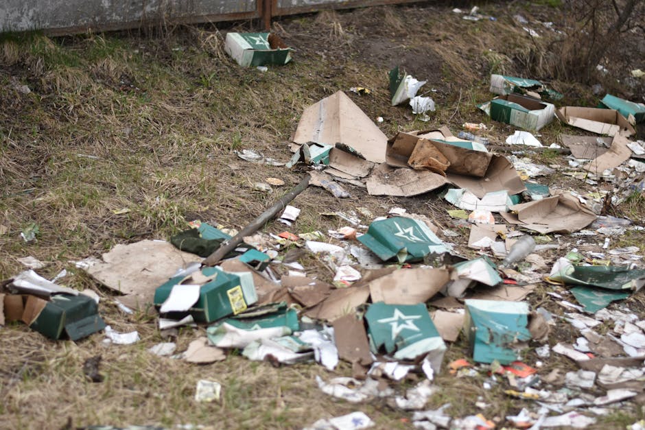An outdoor scene showing multiple overflowing trash and recycling bins placed on a paved sidewalk. The bins contain various waste materials, including paper, cardboard, plastic bags, and packaging, so